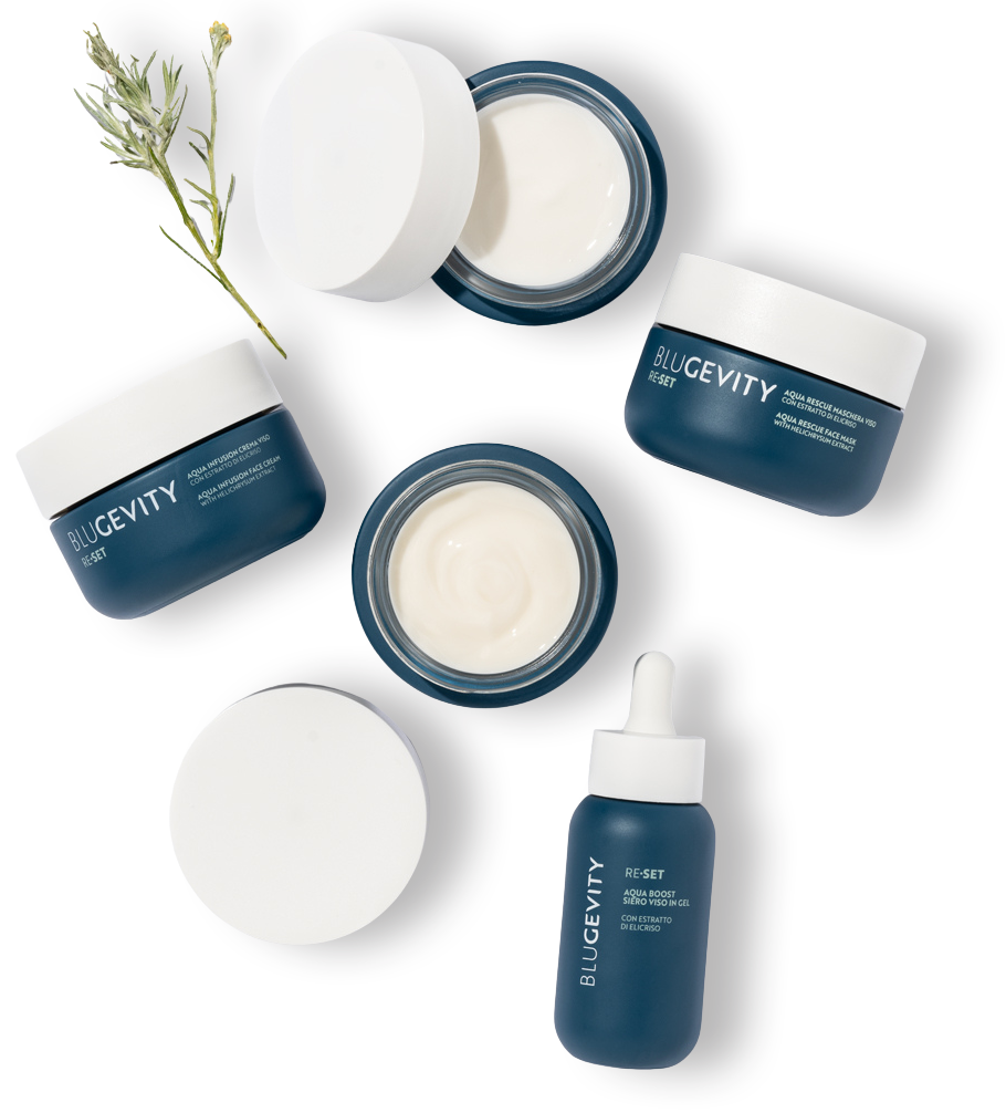 Prodotti per la cura della pelle blu e bianchi disposti su una superficie bianca, con un rametto di verde.