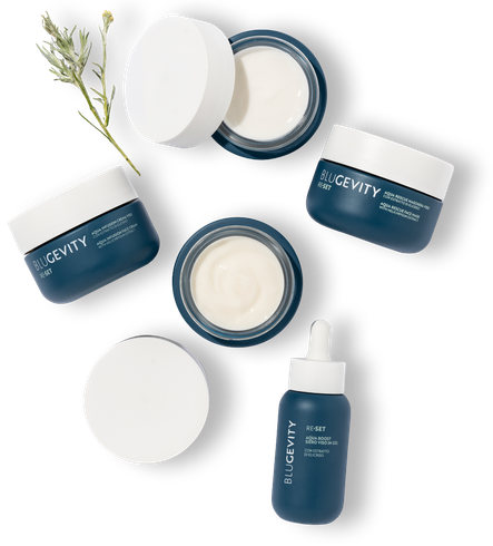 Prodotti per la cura della pelle blu e bianchi disposti su una superficie bianca, con un rametto di verde.