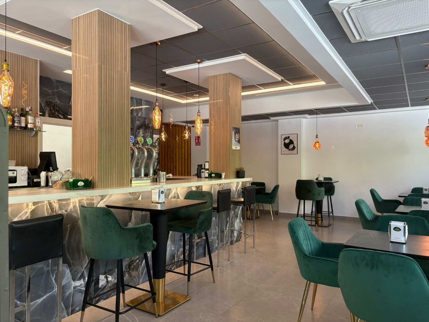 Interior de restaurante con mesas de madera, sillas y barra con botellas de vino y un espejo decorativo.