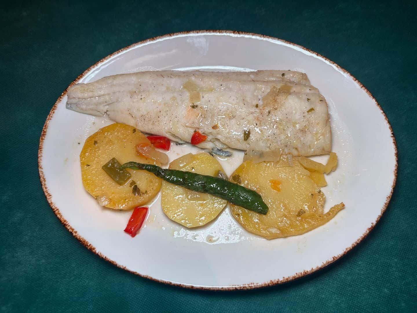 Plato blanco con un trozo de pescado, tres rodajas de patata y pimientos.