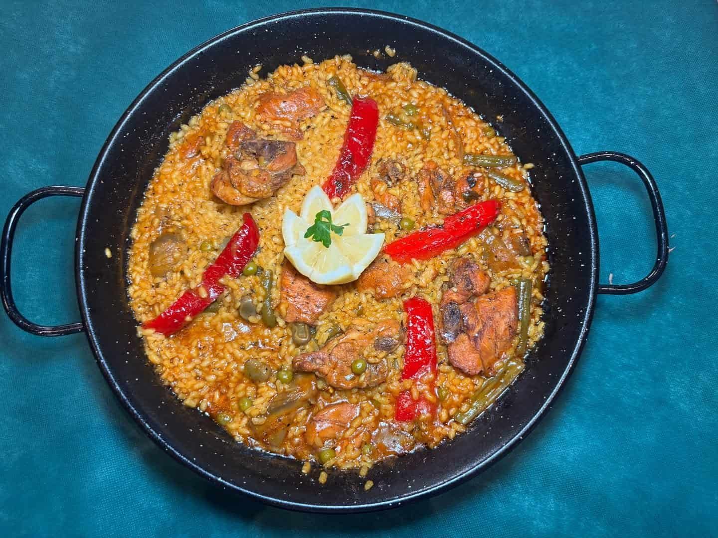Paella en sartén negra, con pollo, arroz, verduras y pimientos rojos; guarnición de limón.