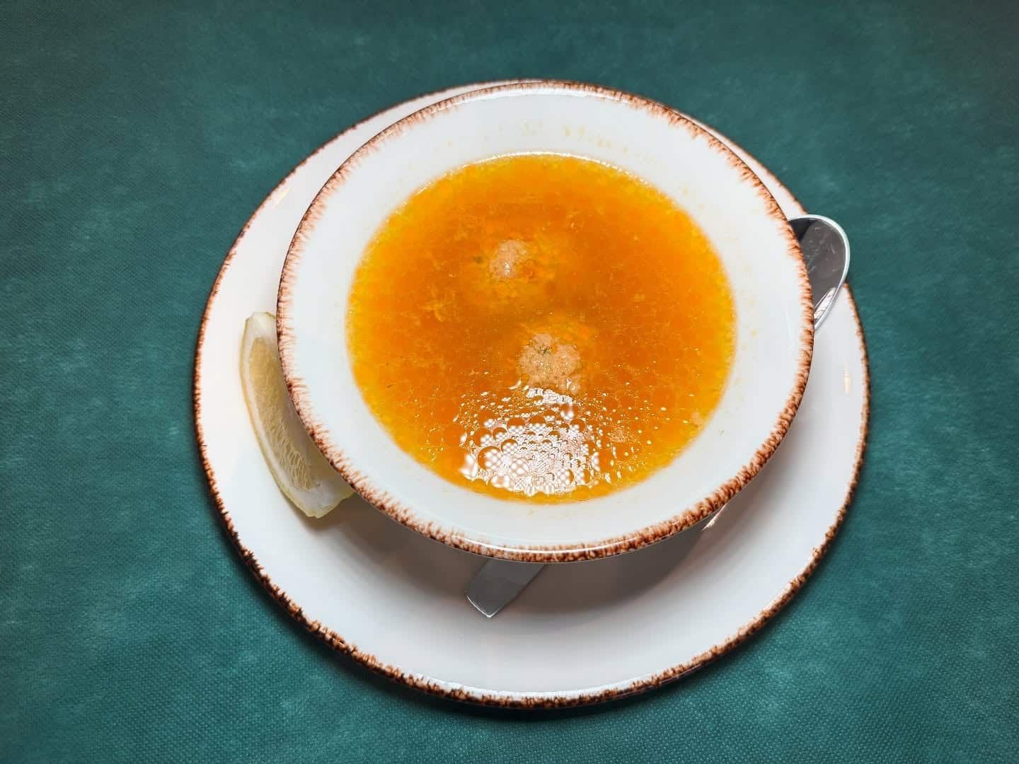 Tazón de sopa de naranja con una rodaja de limón, en un plato blanco, sobre una superficie verde azulado.
