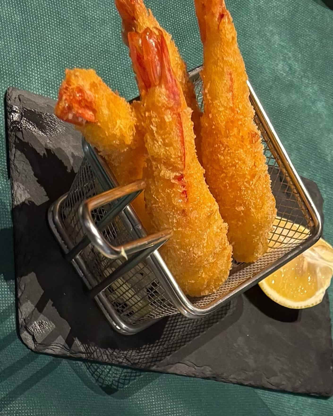 Camarones fritos en una pequeña canasta de metal, servidos en una pizarra con una rodaja de limón.