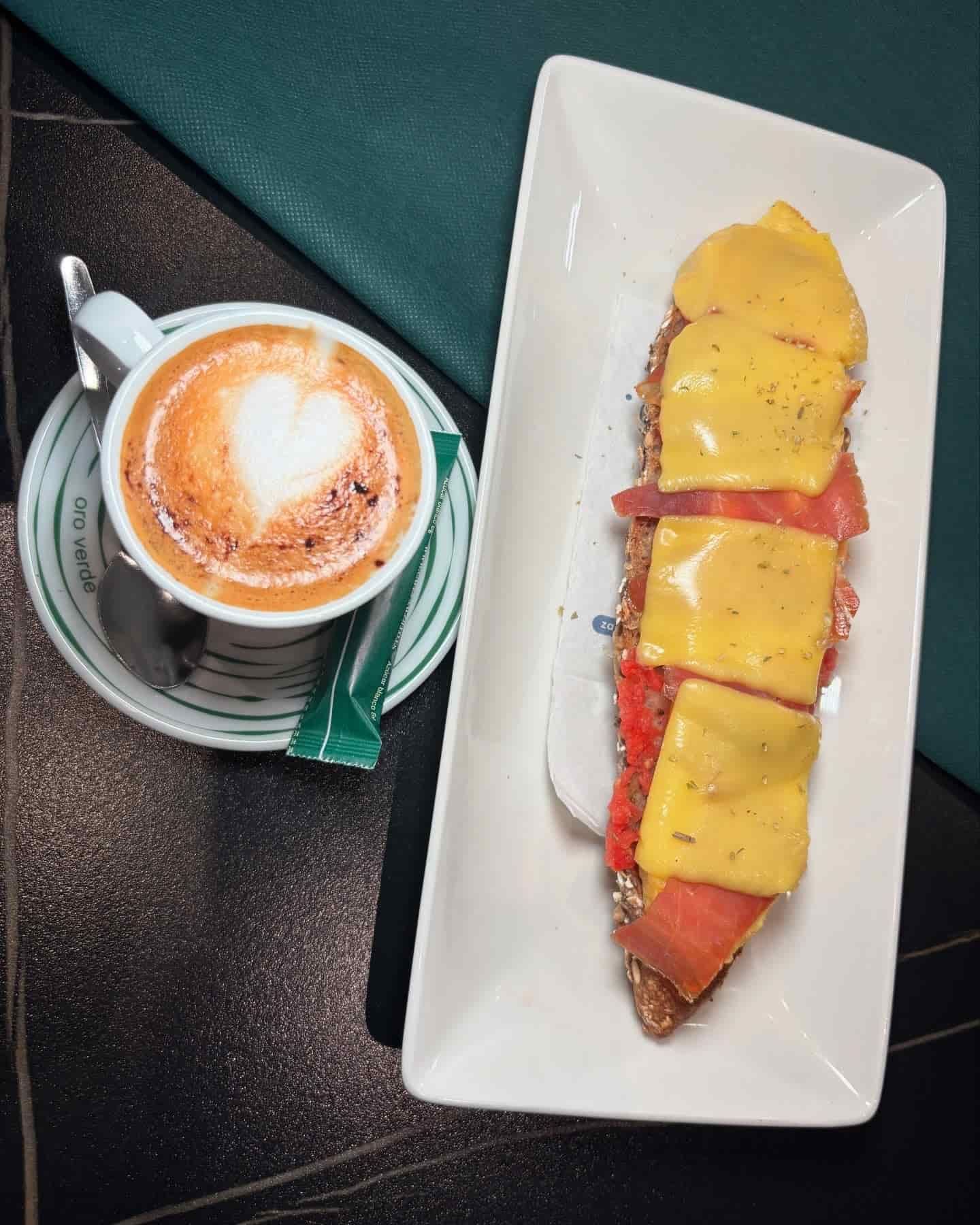 Capuchino con arte de corazón junto a un plato con tostada cubierta con queso y rodajas de tomate.