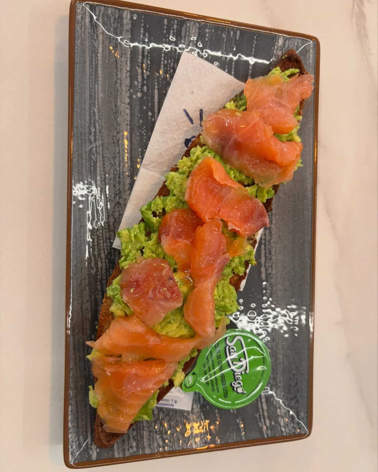 Tostada de aguacate con salmón ahumado en plato rectangular, con paquete de mantequilla y servilleta.