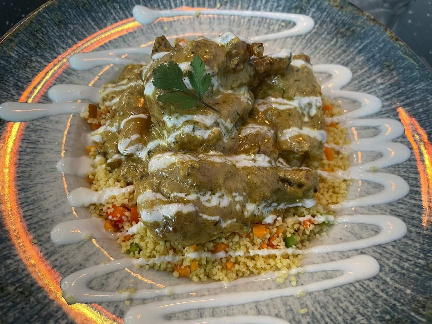 Plato de carne con salsa blanca, servido sobre cuscús con verduras; plato adornado.