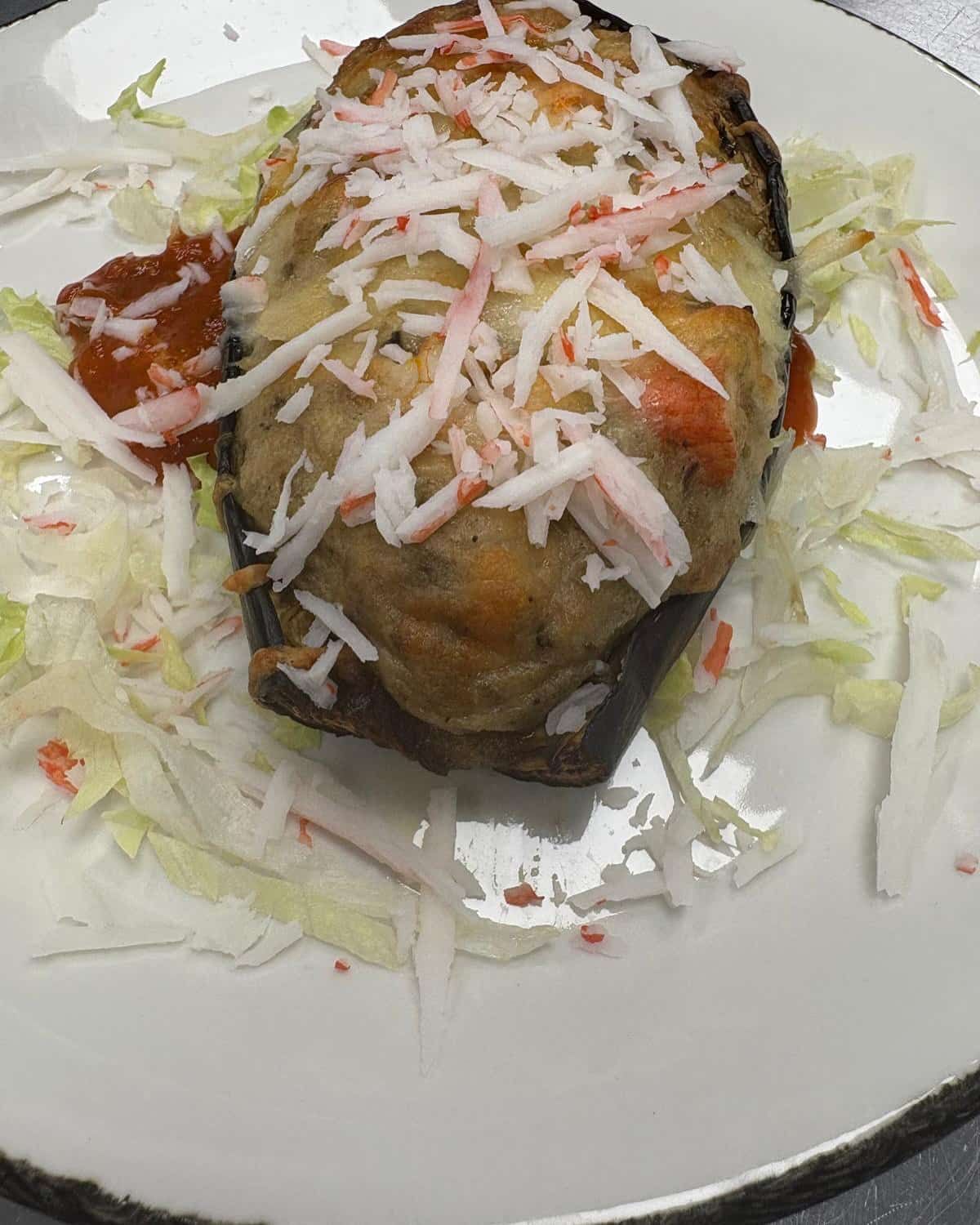 Berenjena rellena al horno, cubierta con cangrejo y salsa, servida sobre lechuga rallada.
