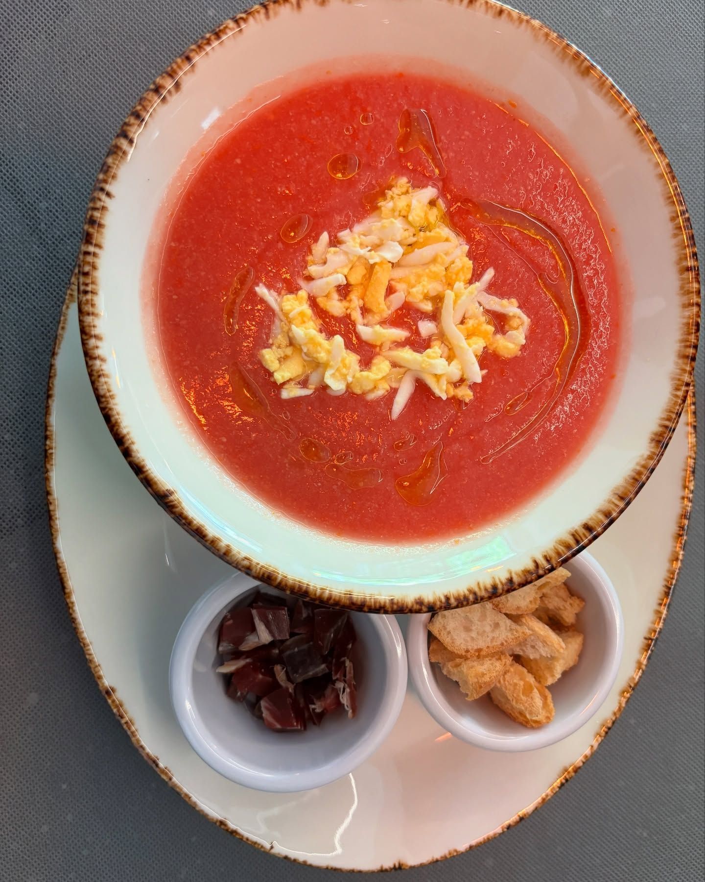 Gazpacho en plato hondo con queso rallado y aceite de oliva, acompañado de jamón y picatostes.