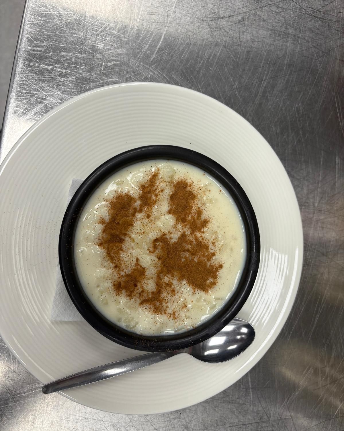 Arroz con leche en un recipiente negro, espolvoreado con canela, sobre un plato blanco con una cuchara.