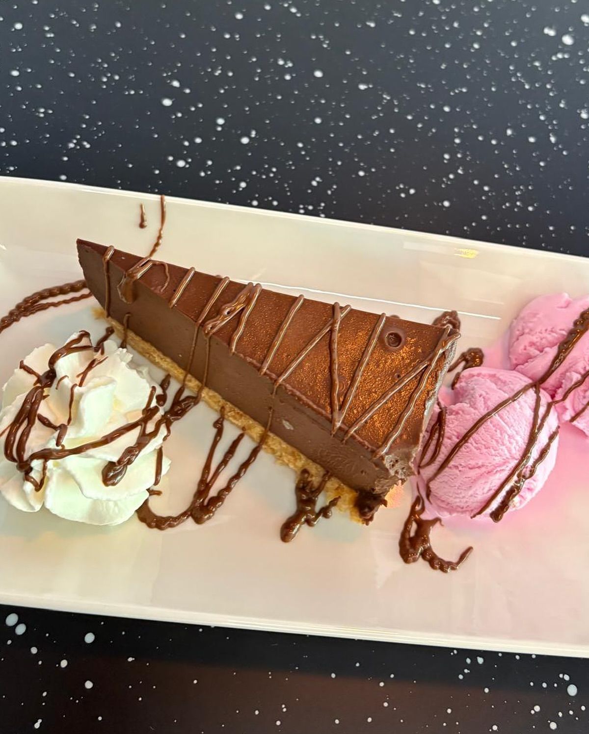 Porción de pastel de chocolate con crema batida y helado rosa, rociado con salsa de chocolate.