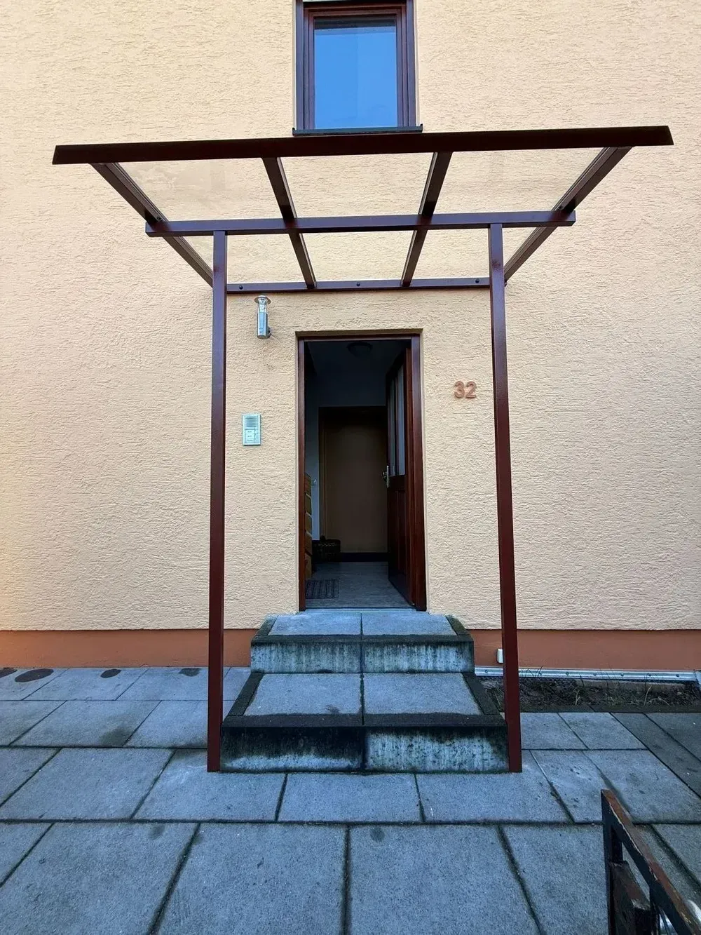 Überdachter Eingang mit Stufen und einem kleinen Vordach vor einer beigefarbenen Gebäudewand.