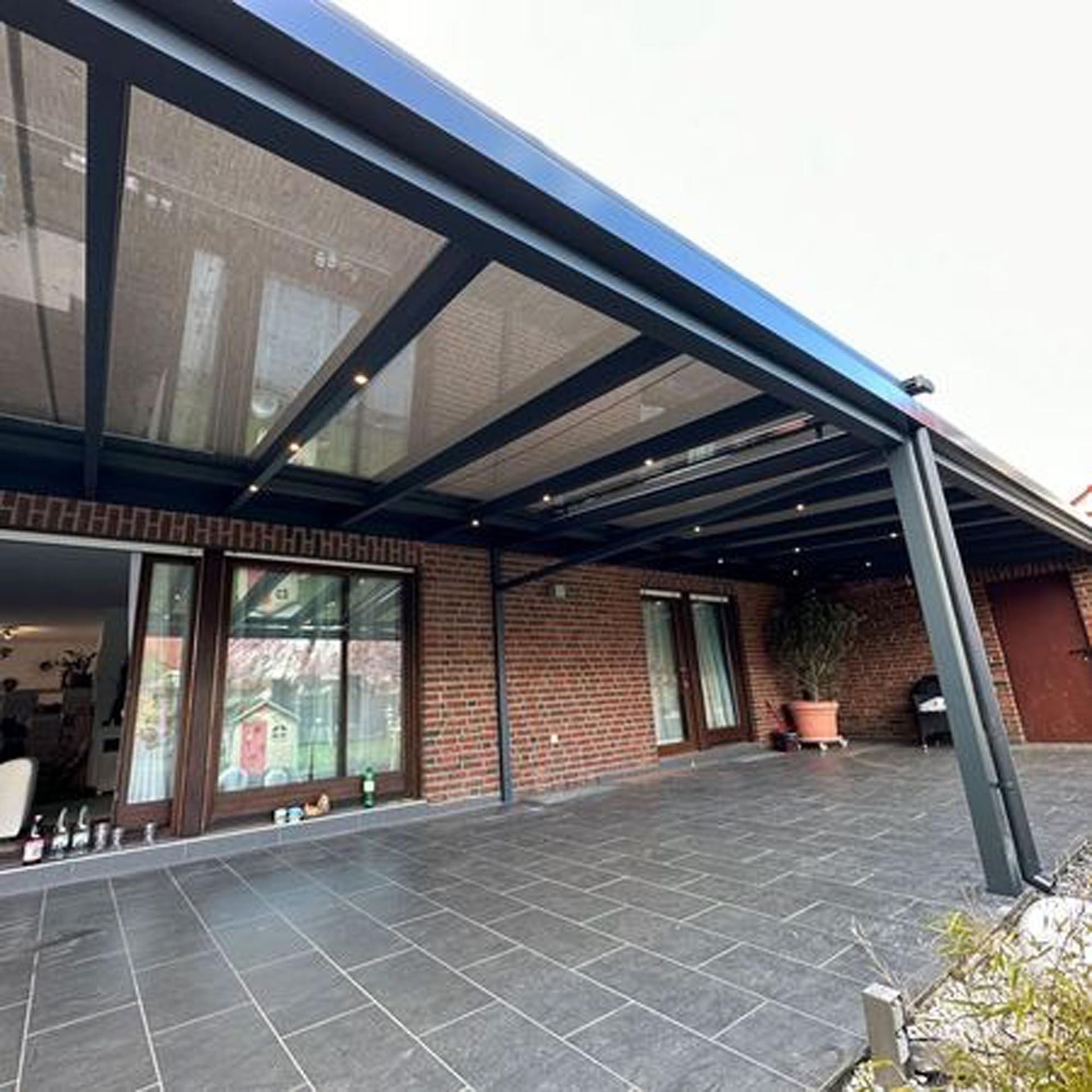 Überdachte Backsteinterrasse mit dunkler Metallmarkise, großem Fliesenboden und Glastüren an der Hausfassade
