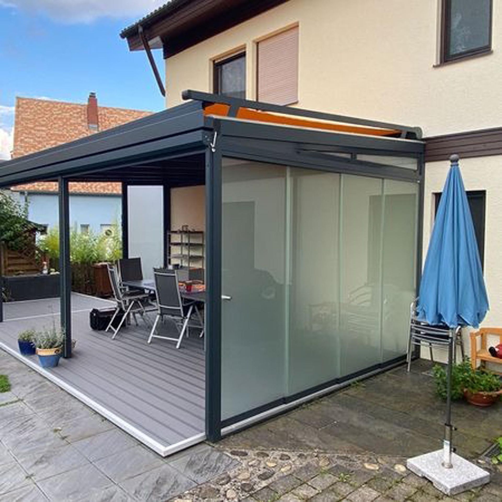 Moderne Terrasse mit dunkel verglaster Überdachung, Holzdeck, Gartentisch, Stühlen und blauem Sonnenschirm