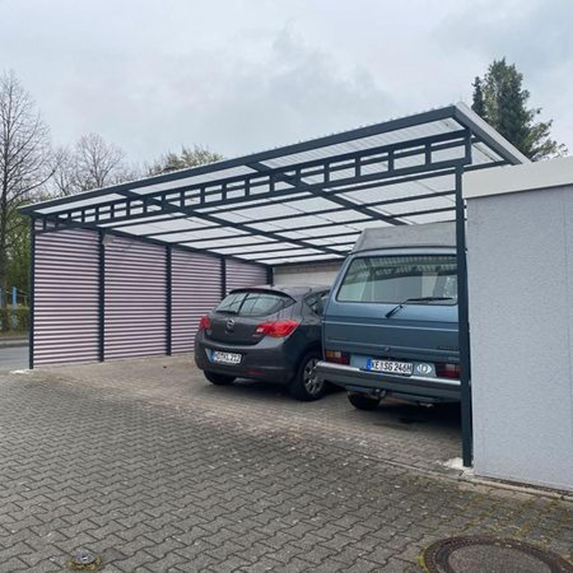 Zwei Autos parken unter einem modernen Carport aus Metall neben einer weißen Mauer.
