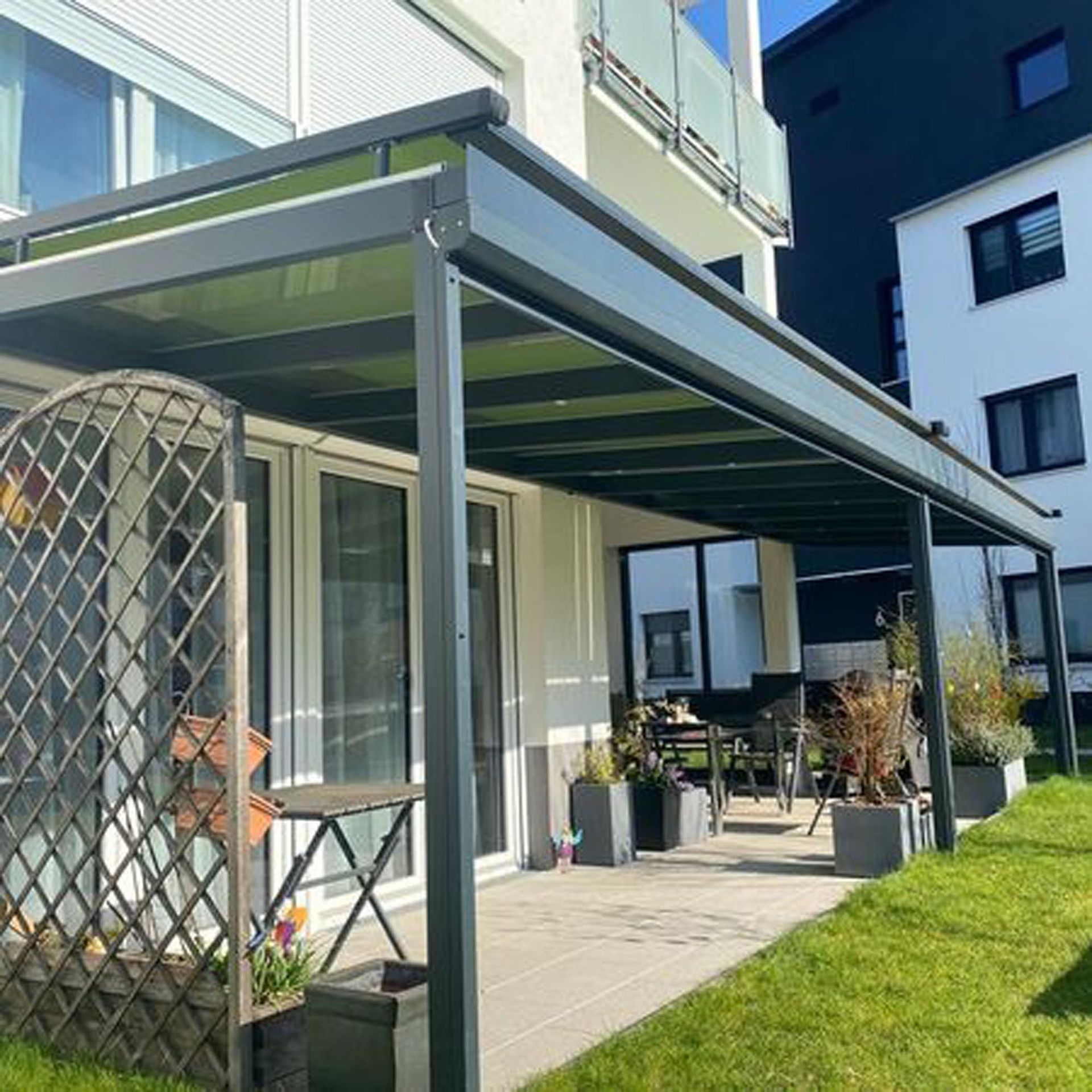 Moderne Terrasse mit grüner Markise, Topfpflanzen und Rasenfläche neben einem Wohngebäude.