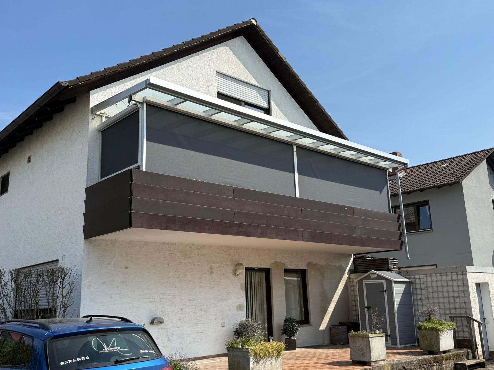 Zweistöckiges weißes Haus mit dunklem Balkon und davor geparktem blauen Auto.