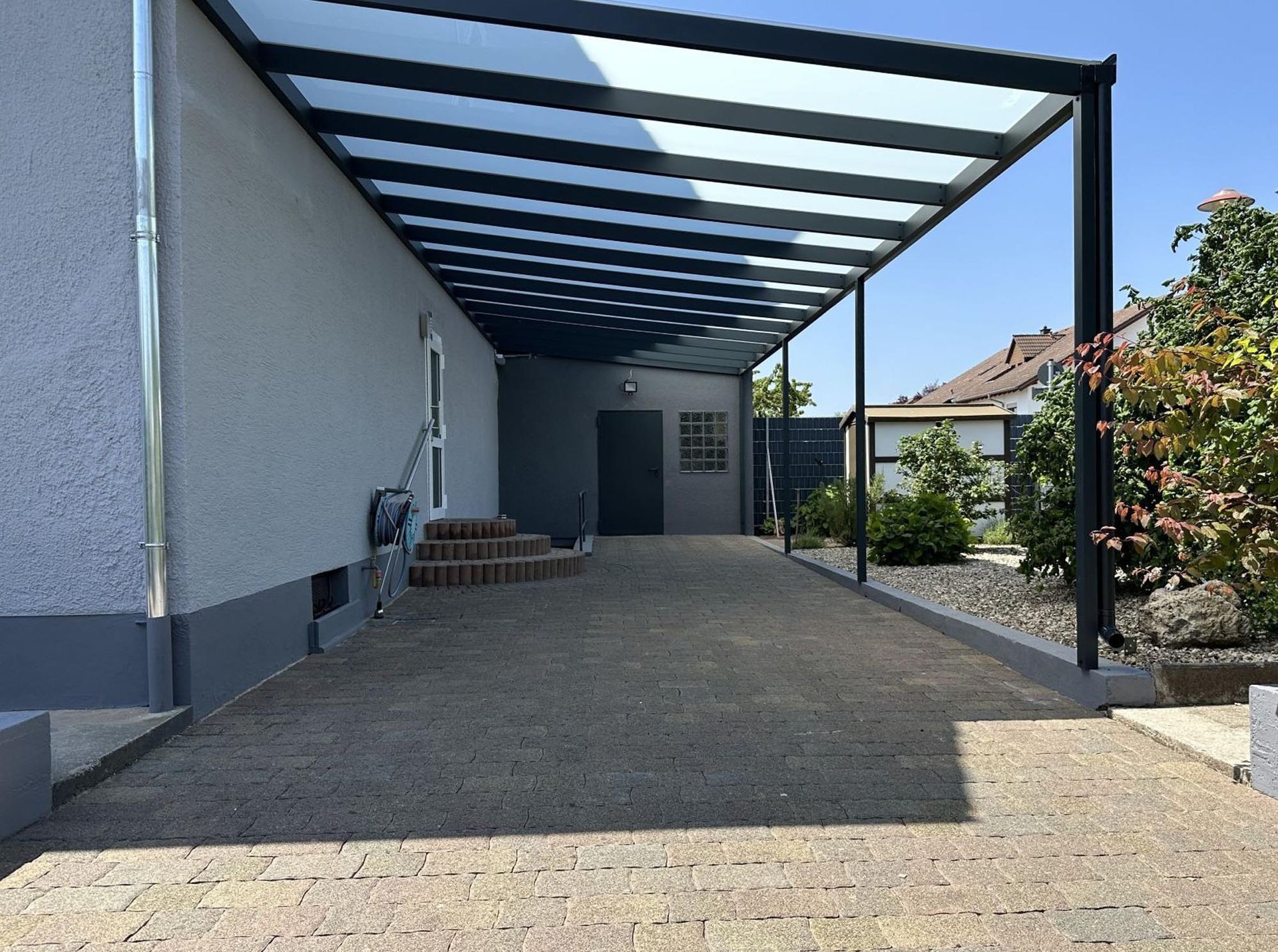 Überdachte Auffahrt neben einem Haus mit Metallvordach und gepflastertem Boden, die zu einer Garage führt