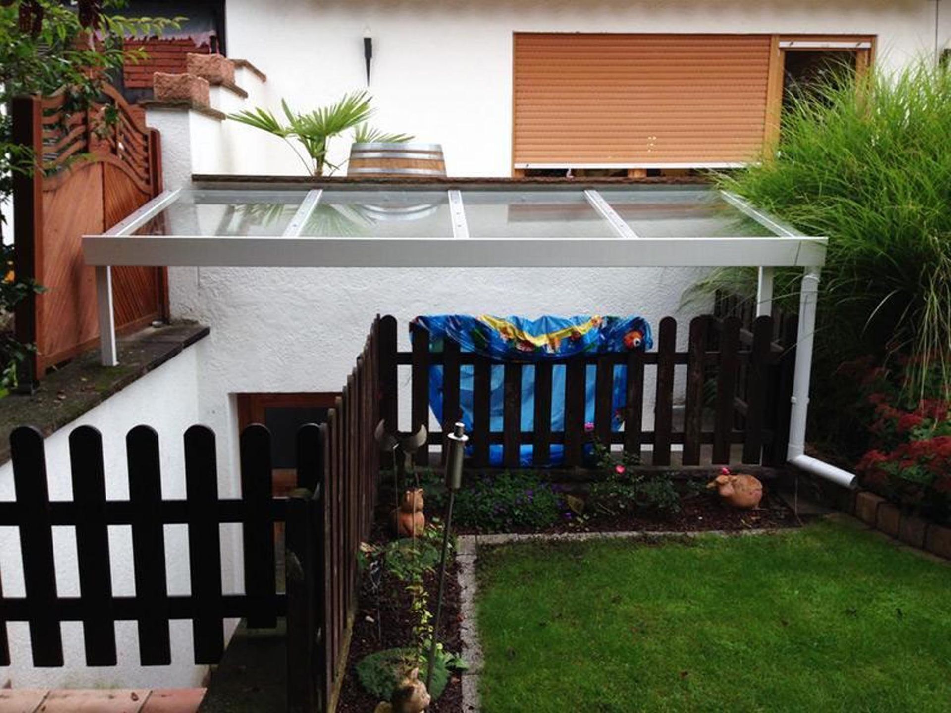 Hinterhofterrasse mit weißer Pergola, schwarzem Zaun, grünem Rasen und dichter Bepflanzung an den Rändern