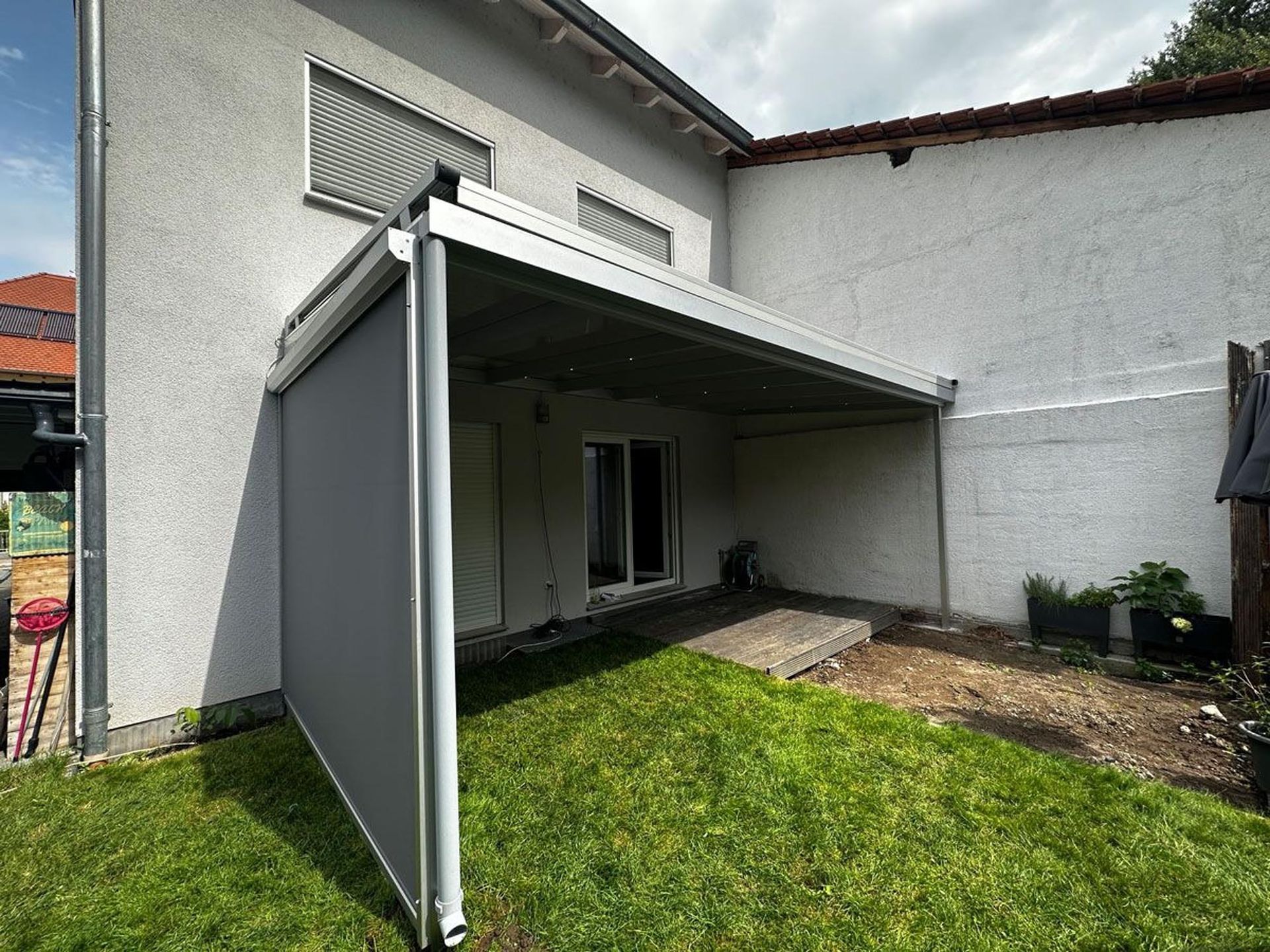 Eine mit einer grauen Markise überdachte Terrasse steht neben einem weißen Haus, im Vordergrund ist ein grüner Rasen zu sehen.