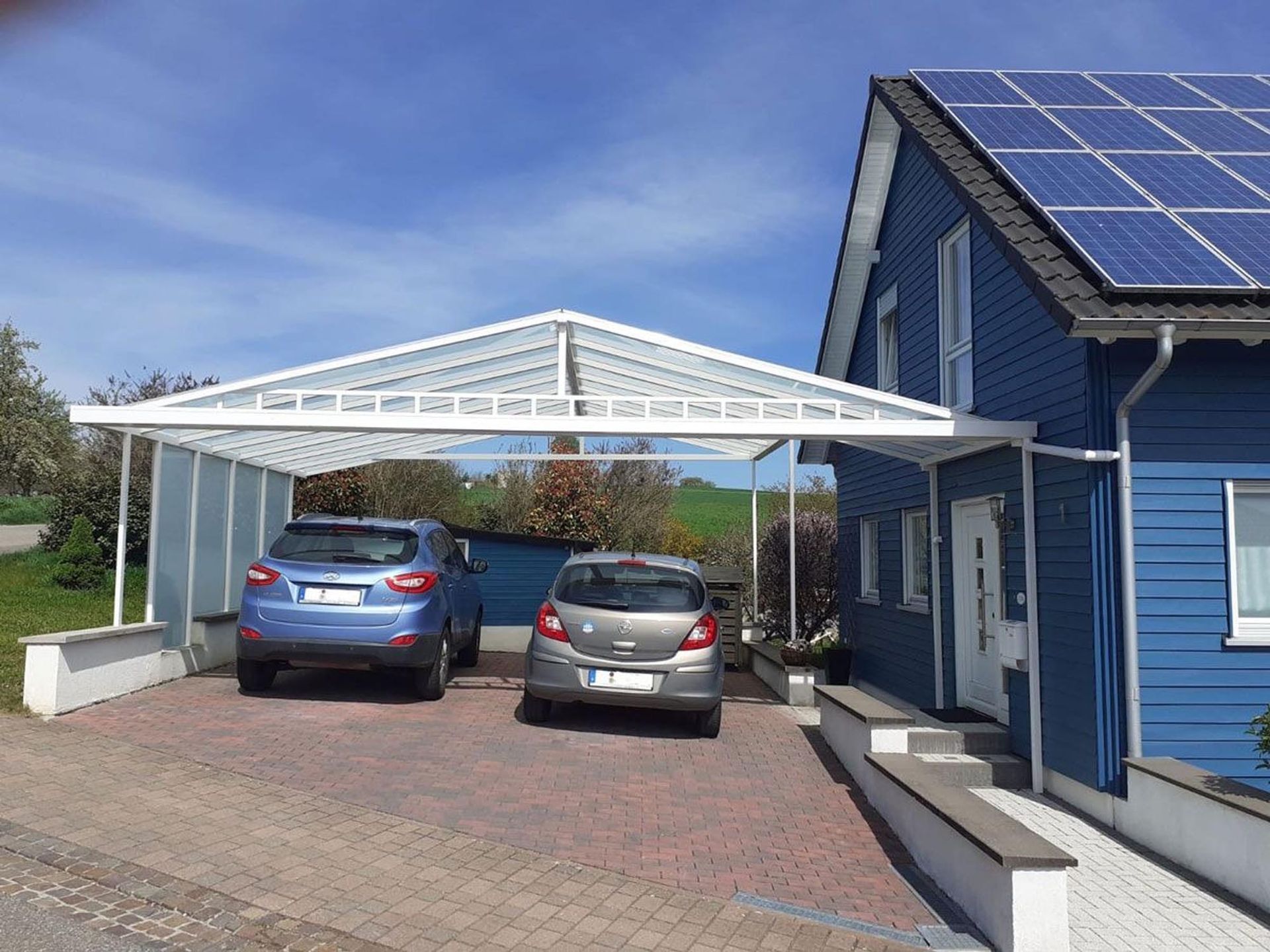 Zwei Autos parken unter einem weißen Carport neben einem blauen Haus mit Solarpaneelen.