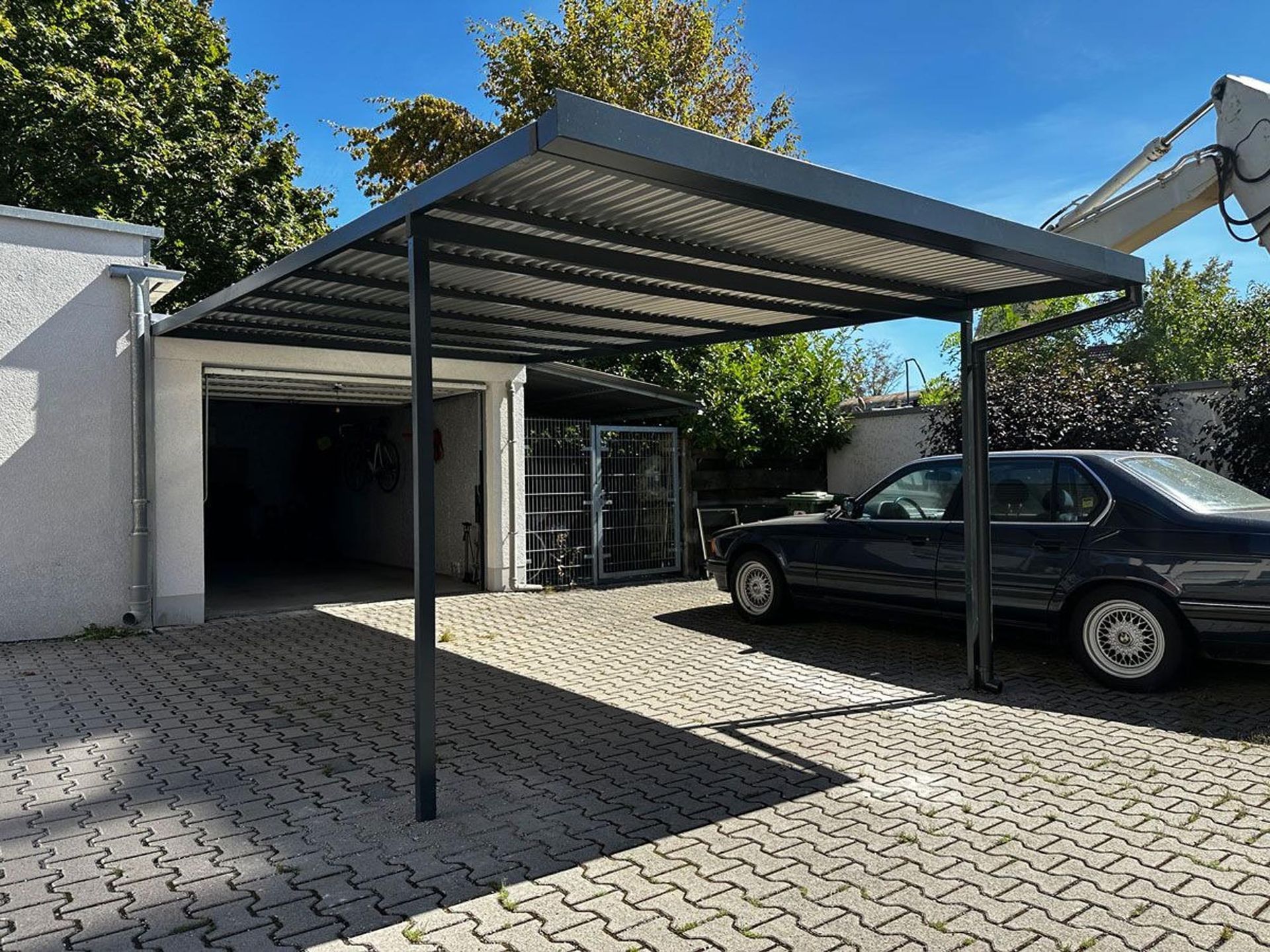 Ein Carport mit einem schwarzen Auto, das neben einer Garage auf einer gepflasterten Auffahrt geparkt ist.