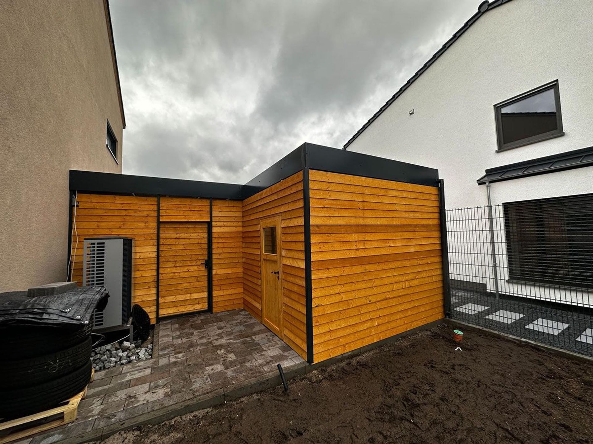 Ein mit Holz getäfelter Terrassenschuppen in einem schmalen Hof, darüber graue Pflastersteine ​​und bewölkter Himmel.