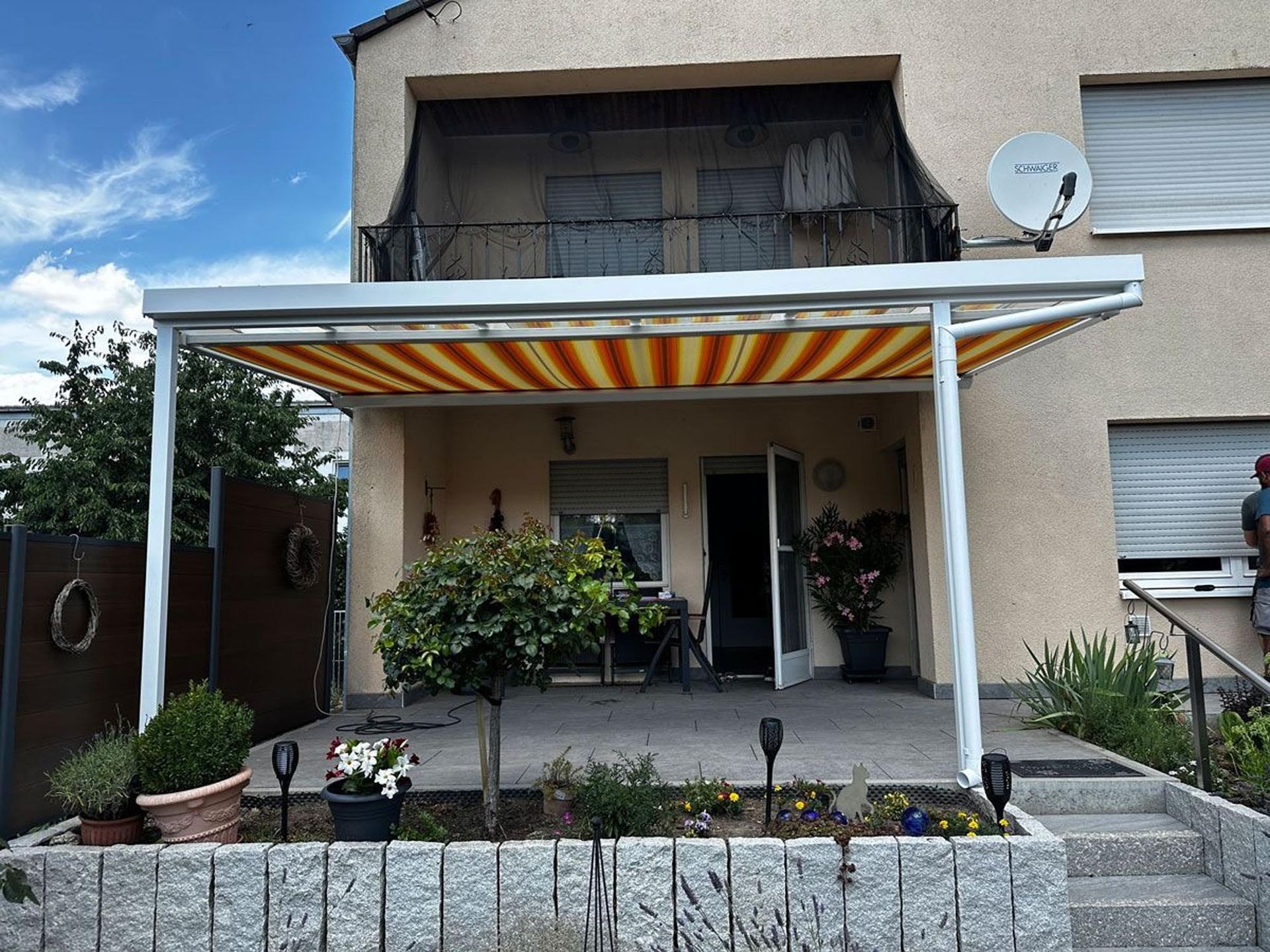Zweistöckiges Haus mit gestreifter Markise, Veranda, Treppe, Topfpflanzen und Satellitenschüssel.