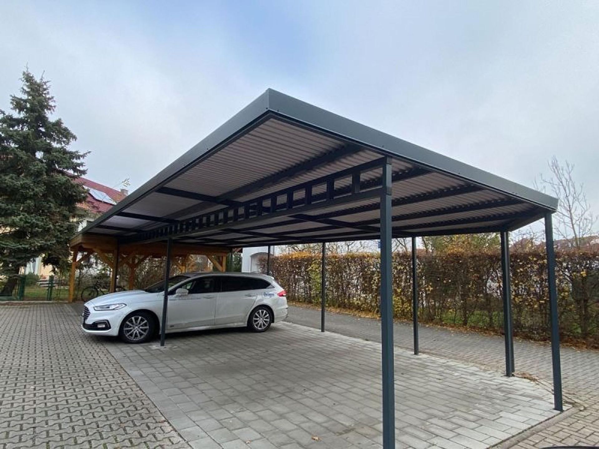 Ein silberner Pkw parkte unter einem Carport aus Metall in einer gepflasterten Auffahrt, dahinter befanden sich Hecken und Bäume.