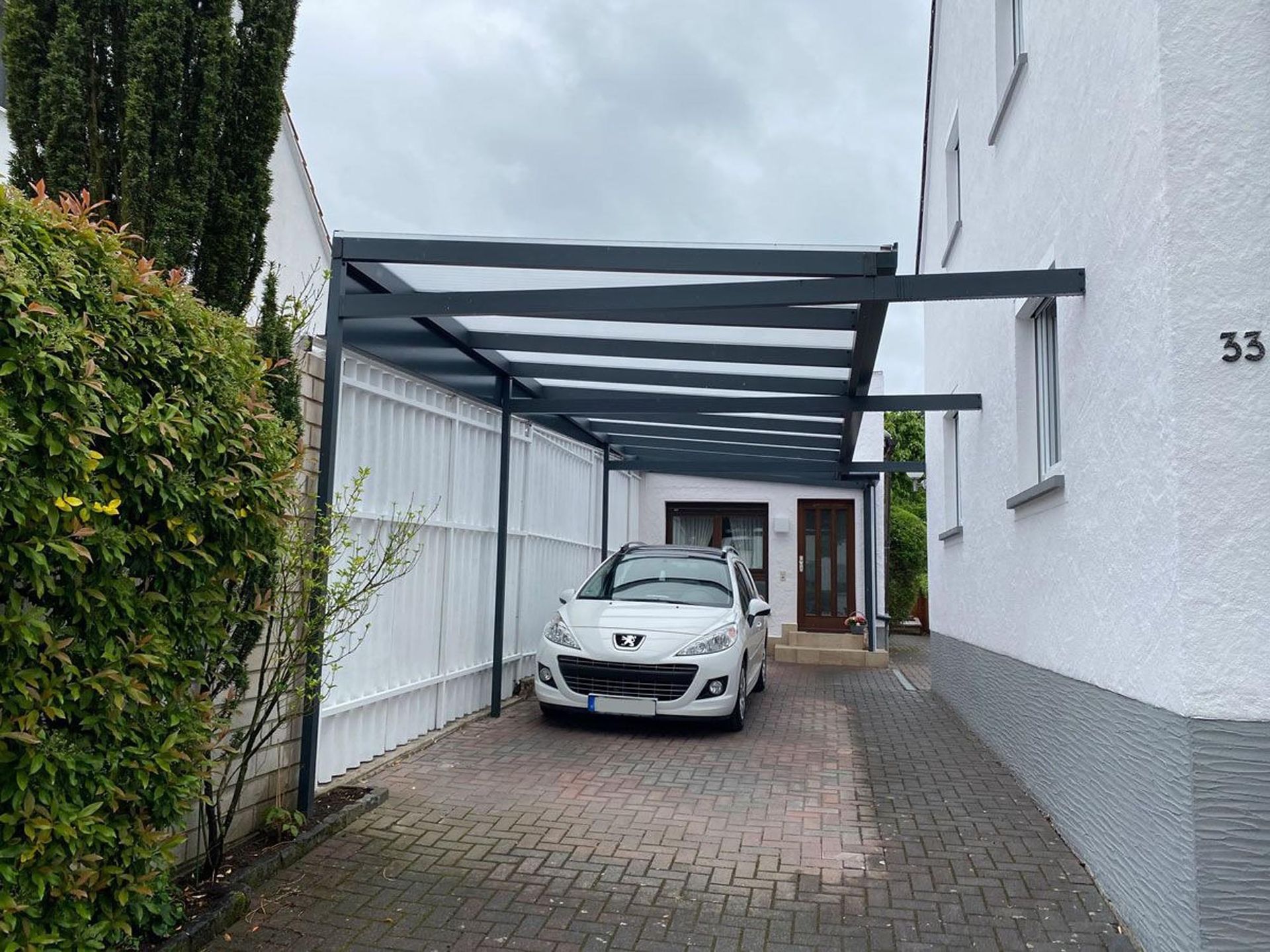 Auffahrt mit einem weißen Auto, das unter einem Carport aus Metall neben weißen Gebäuden geparkt ist.