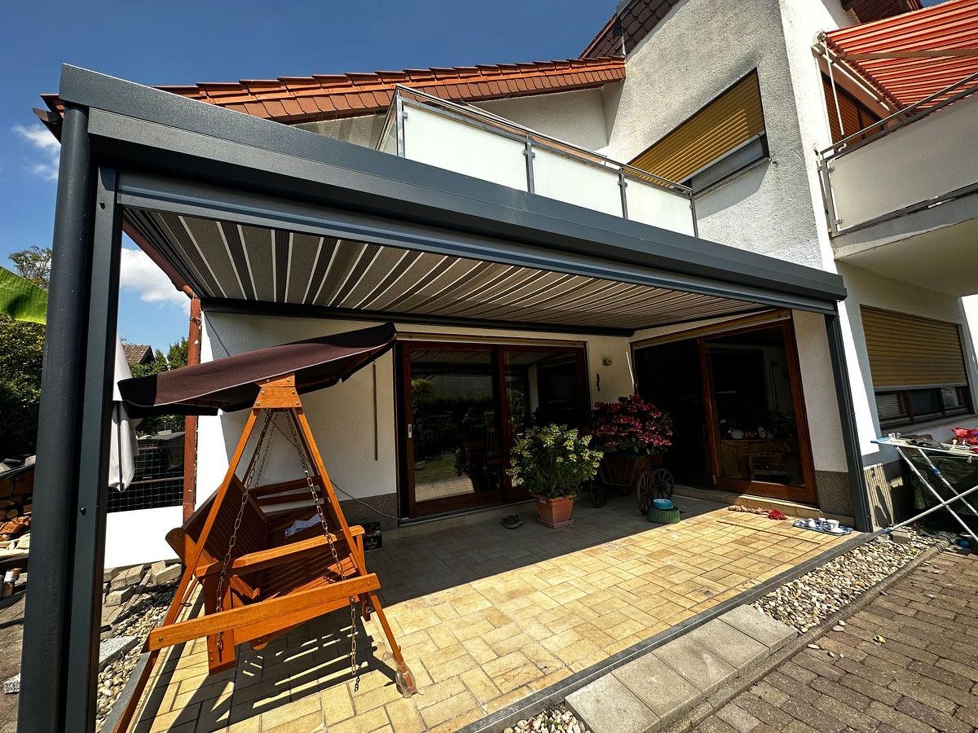 Terrasse mit großer grauer Markise, hölzerner Schaukelstuhl, Topfpflanzen und einer Hausfassade im hellen Sonnenlicht