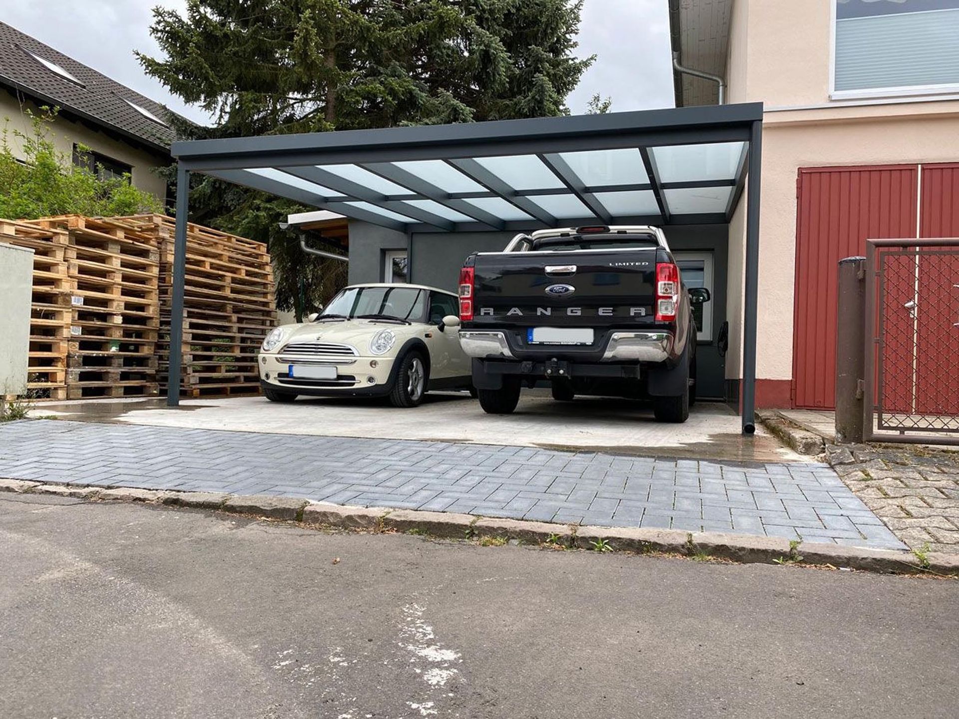 Zwei Autos parken unter einem gläsernen Carport neben einem Haus und einem roten Tor.