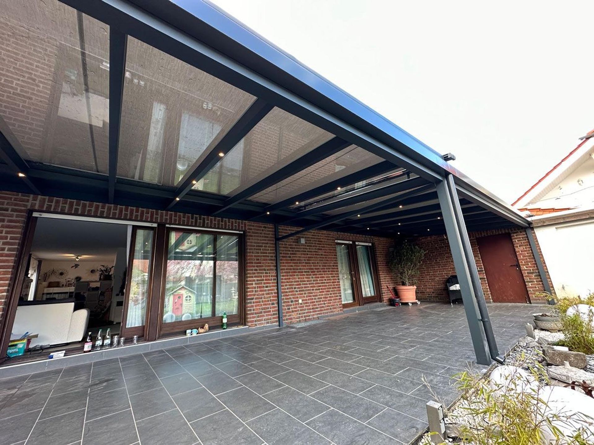 Überdachte Ziegelterrasse mit schwarzer Metallpergola und Fliesenboden neben einem Haus