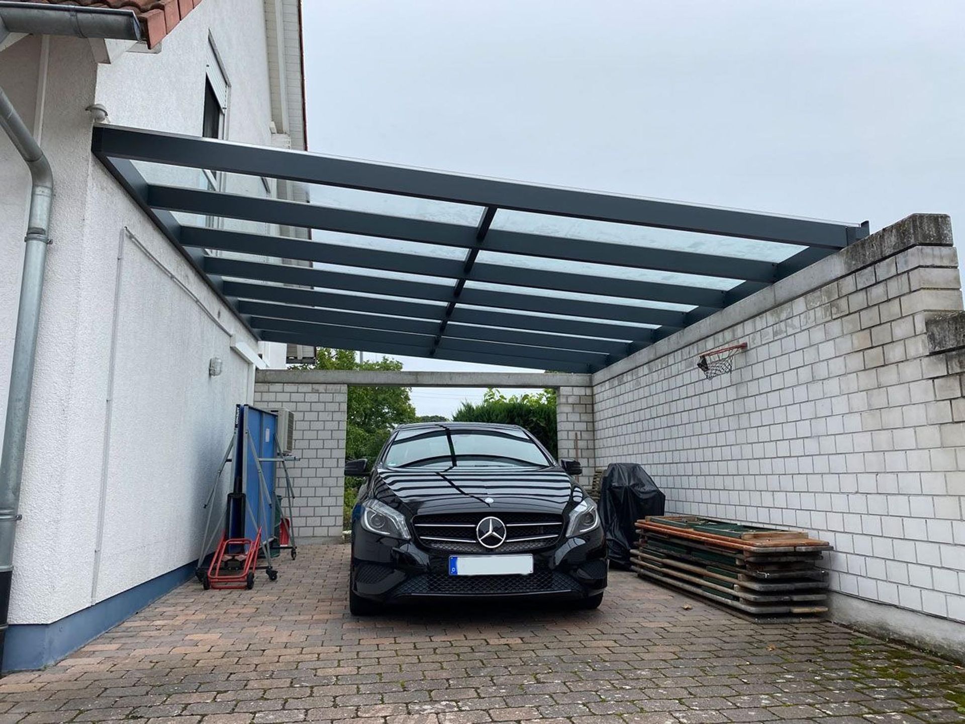 Ein schwarzes Auto parkte unter einem Carport aus Metall neben weißen Backsteinmauern mit gestapelten Materialien.