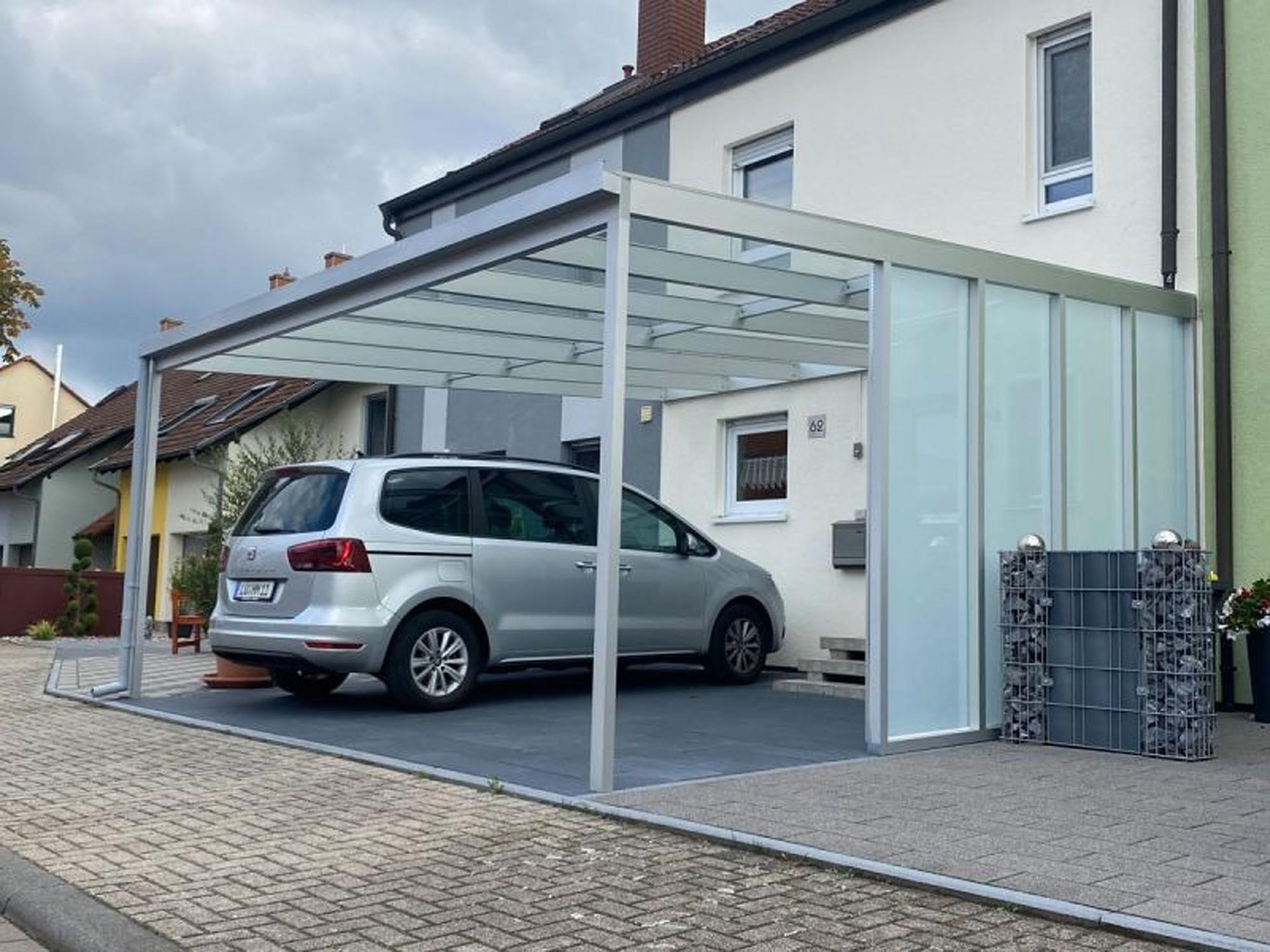 Ein silberner Minivan parkt unter einem modernen Carport aus Glas neben einem weißen Haus.