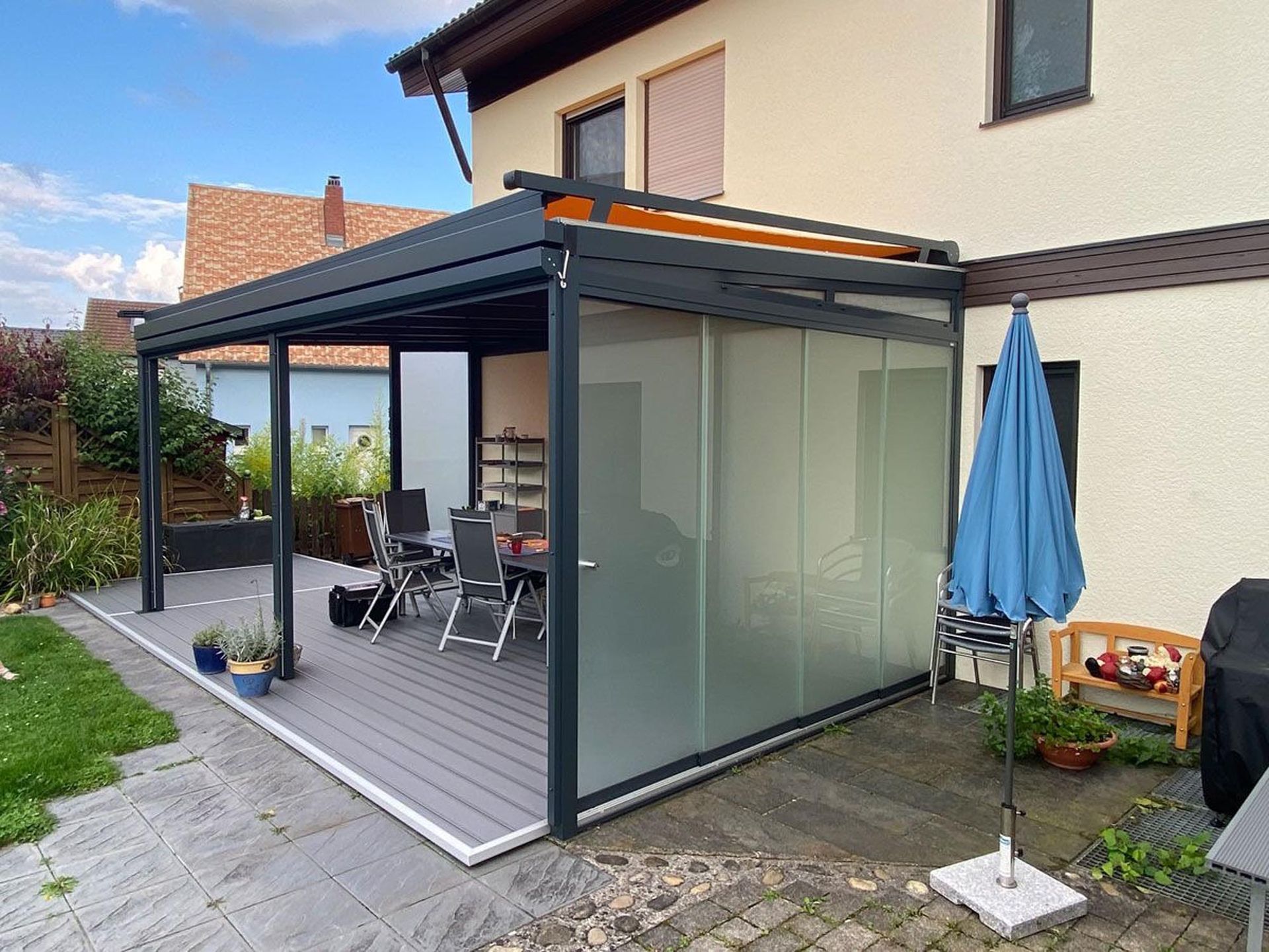 Moderne Terrasse mit schwarz umrandeter Glasfassade, grauem Terrassenbelag, Gartenmöbeln und einem blauen Sonnenschirm neben einem Haus