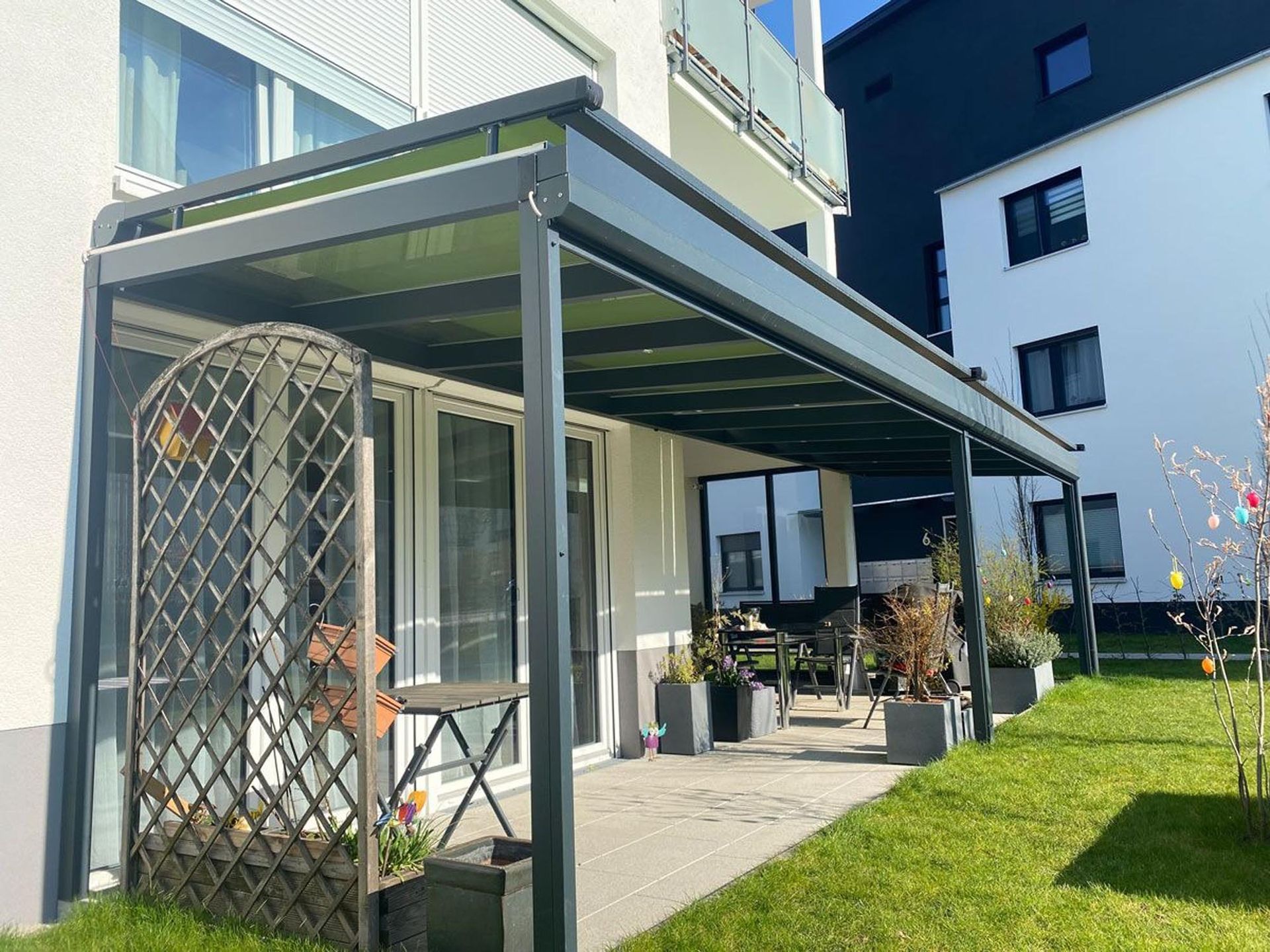 Moderne Hausterrasse mit überdachter Pergola, Tisch und Stühlen sowie einem grünen Rasen unter klarem Himmel.