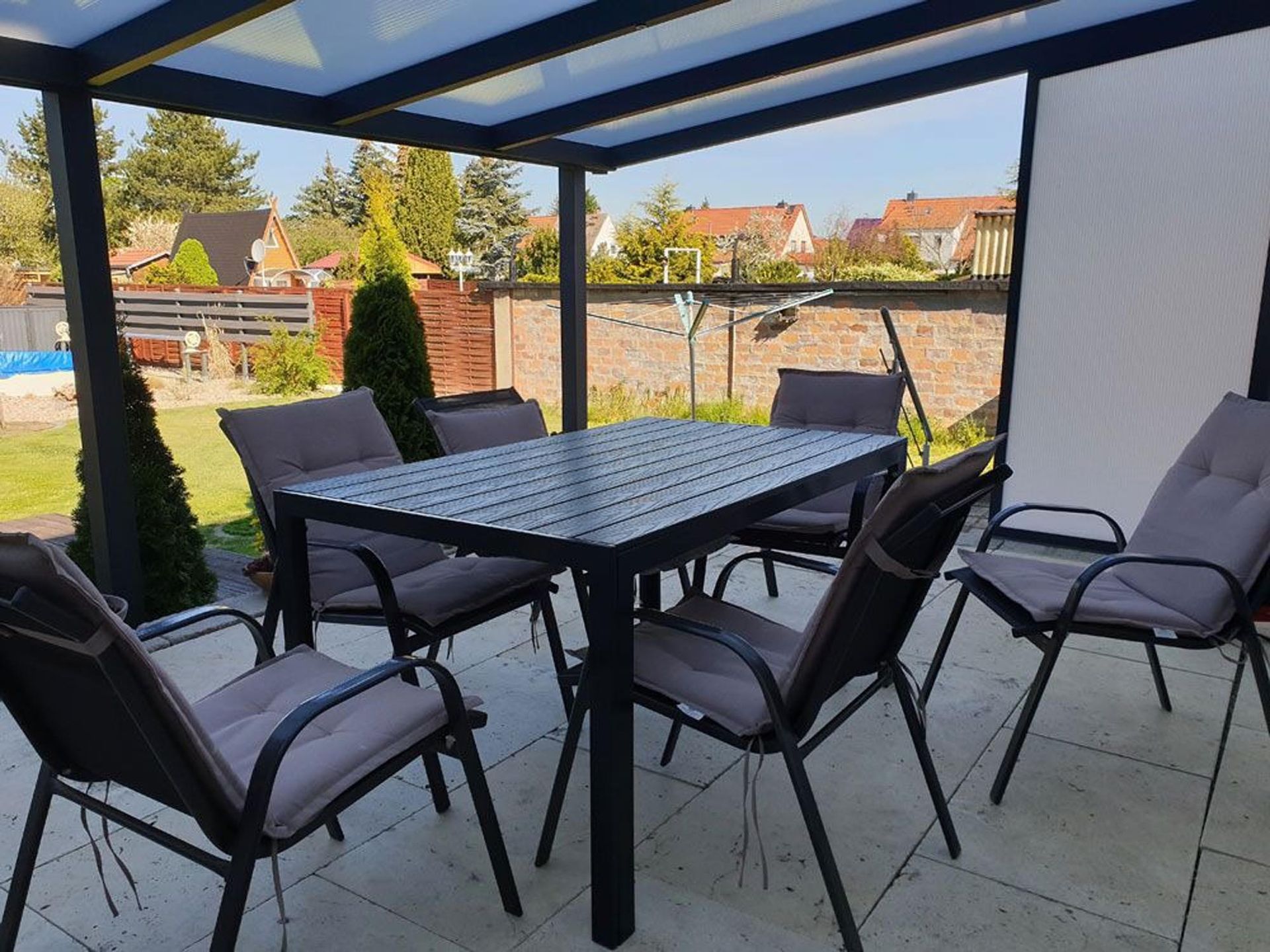 Überdachte Terrasse mit schwarzem Metalltisch und Stühlen, mit Blick auf eine Backsteinmauer und die dahinterliegenden Häuser.