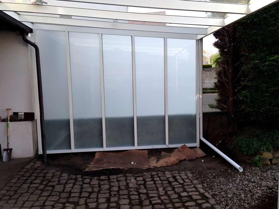 Weiße, mattierte Glasschiebetüren unter einer überdachten Terrasse neben einem gepflasterten Gehweg und einer Hecke