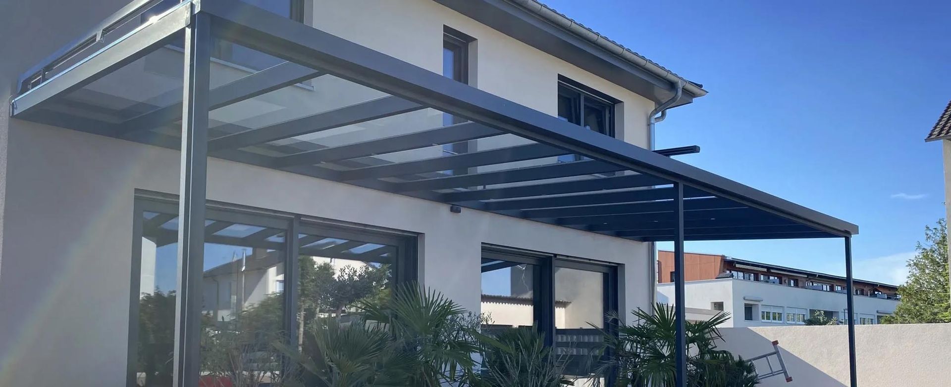 Überdachte Terrasse mit grauem Metallrahmen, an ein Haus angebaut, unter klarem, blauem Himmel