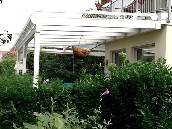 Weiße Terrassenpergola über üppigen grünen Sträuchern mit einem Hängekorb darunter
