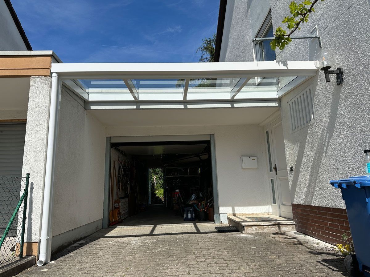 Weiße Garageneinfahrt neben einem Haus, mit einem Carportdach aus Glas und einem blauen Recyclingbehälter auf der rechten Seite.