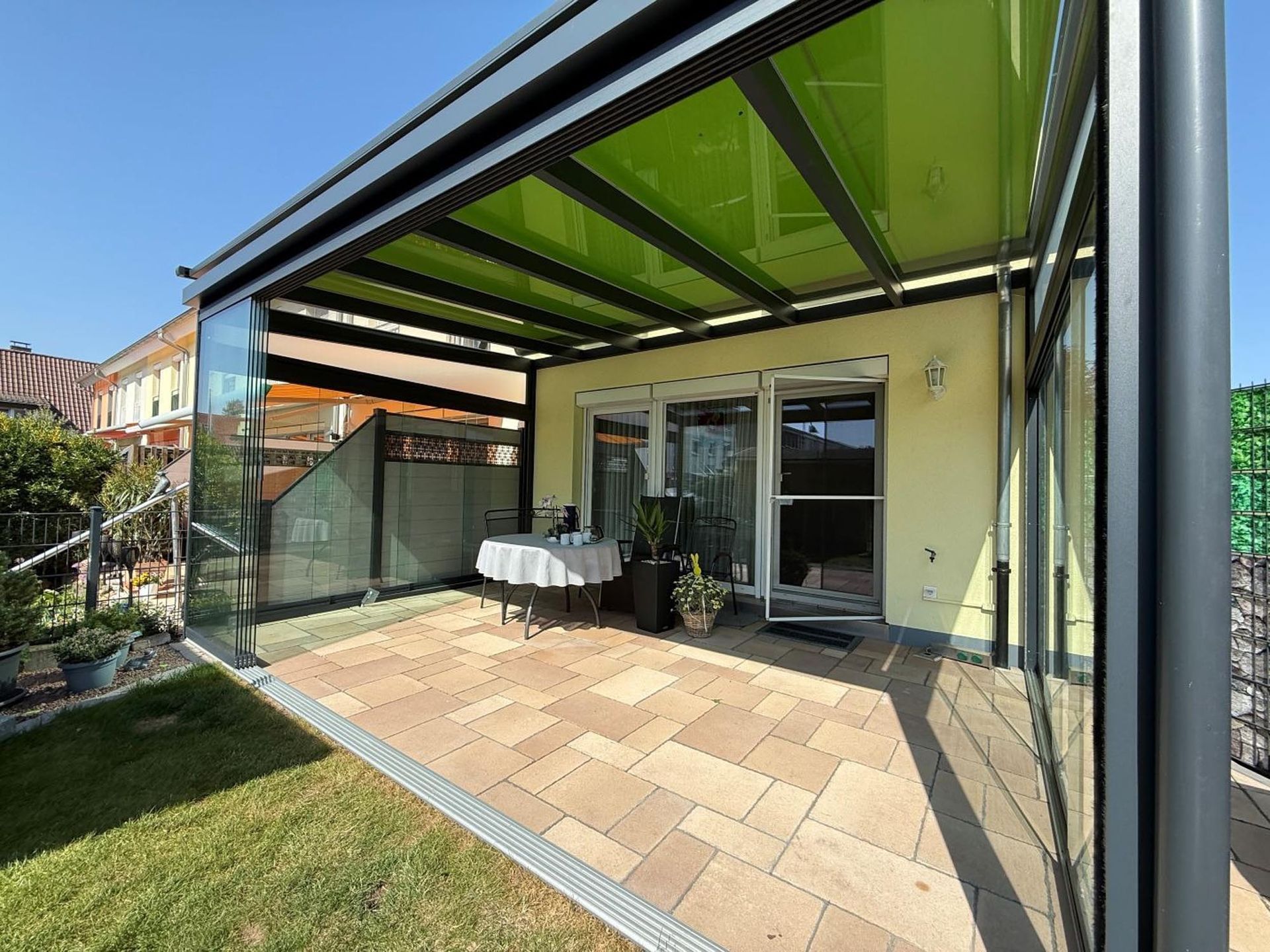 Moderne Terrasse mit Glastüren, schwarzer Markise, Fliesenboden und einem kleinen Tisch neben einem Rasengarten