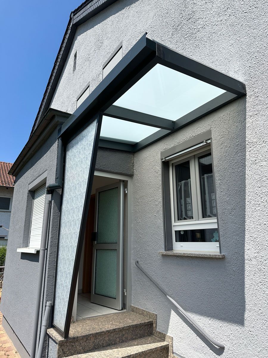 Hauseingang mit grauen Stuckwänden, einer grünen Tür und einem Glasvordach über den Stufen