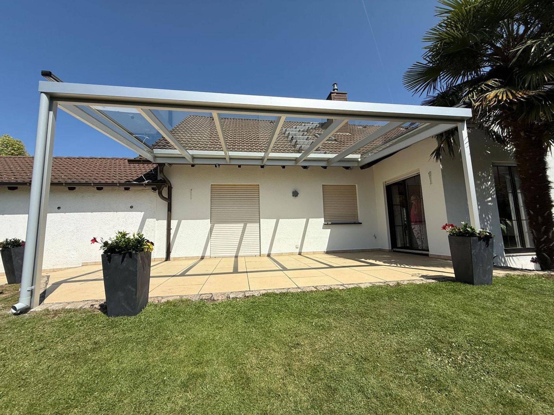 Überdachte Terrasse mit weißen Balken und Ziegeldach über einem Rasen, flankiert von zwei schwarzen Pflanzkästen.