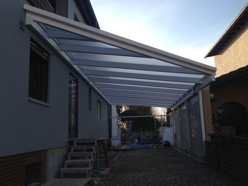 Ein Carport aus Metall mit lichtdurchlässigen Dachplatten steht neben einem Haus und überspannt eine Kiesauffahrt.