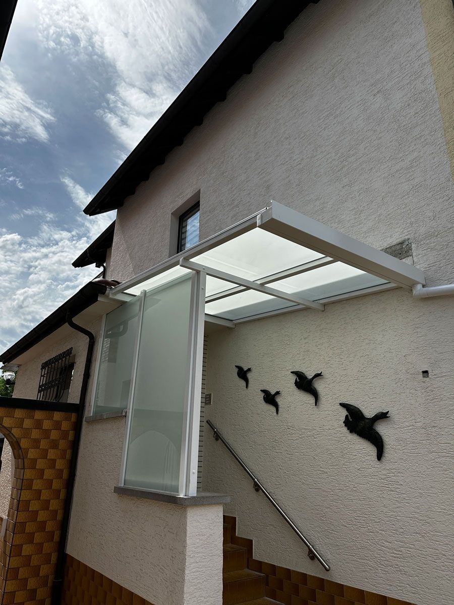 Vordereingang eines Hauses mit Glasvordach, grauer Stuckwand und Vogelsilhouetten auf den Stufen.