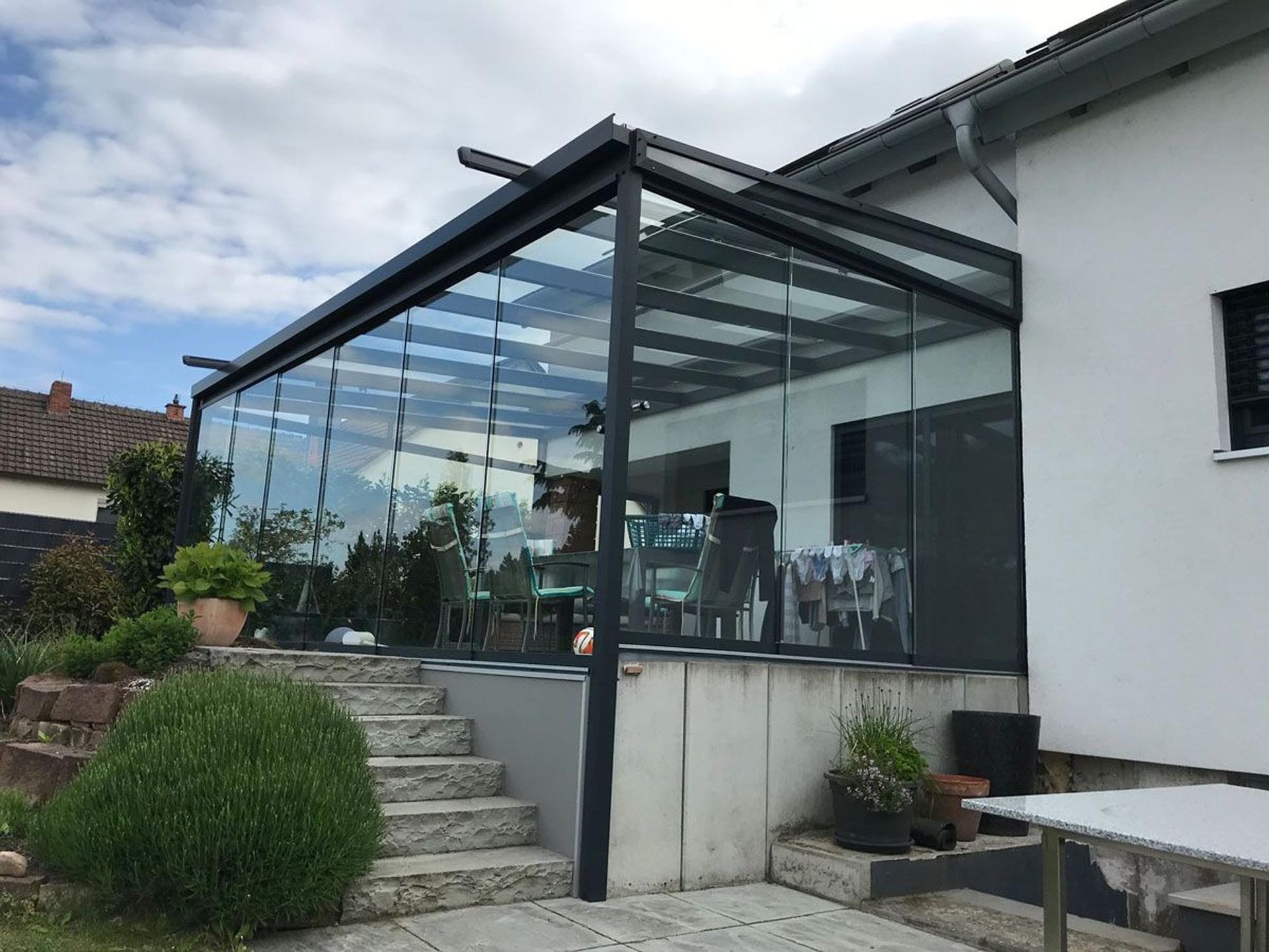 Verglaste Terrasse mit schwarzem Rahmen neben einem weißen Haus, die zu Steinstufen und Gartenpflanzen führt.