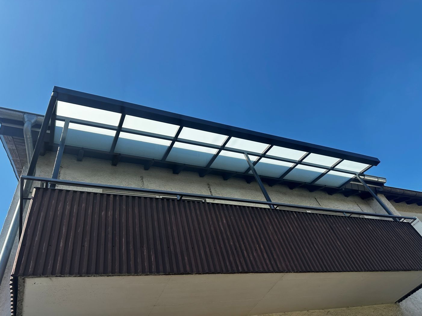 Gebäudefassade mit dunklen, vertikalen Lamellen und einem lichtdurchlässigen Dachvordach vor blauem Himmel