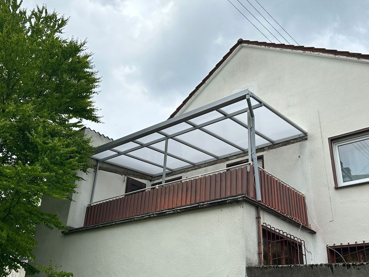 Balkon eines weißen Hauses mit lichtdurchlässiger Metallmarkise und braunem Geländer unter bewölktem Himmel