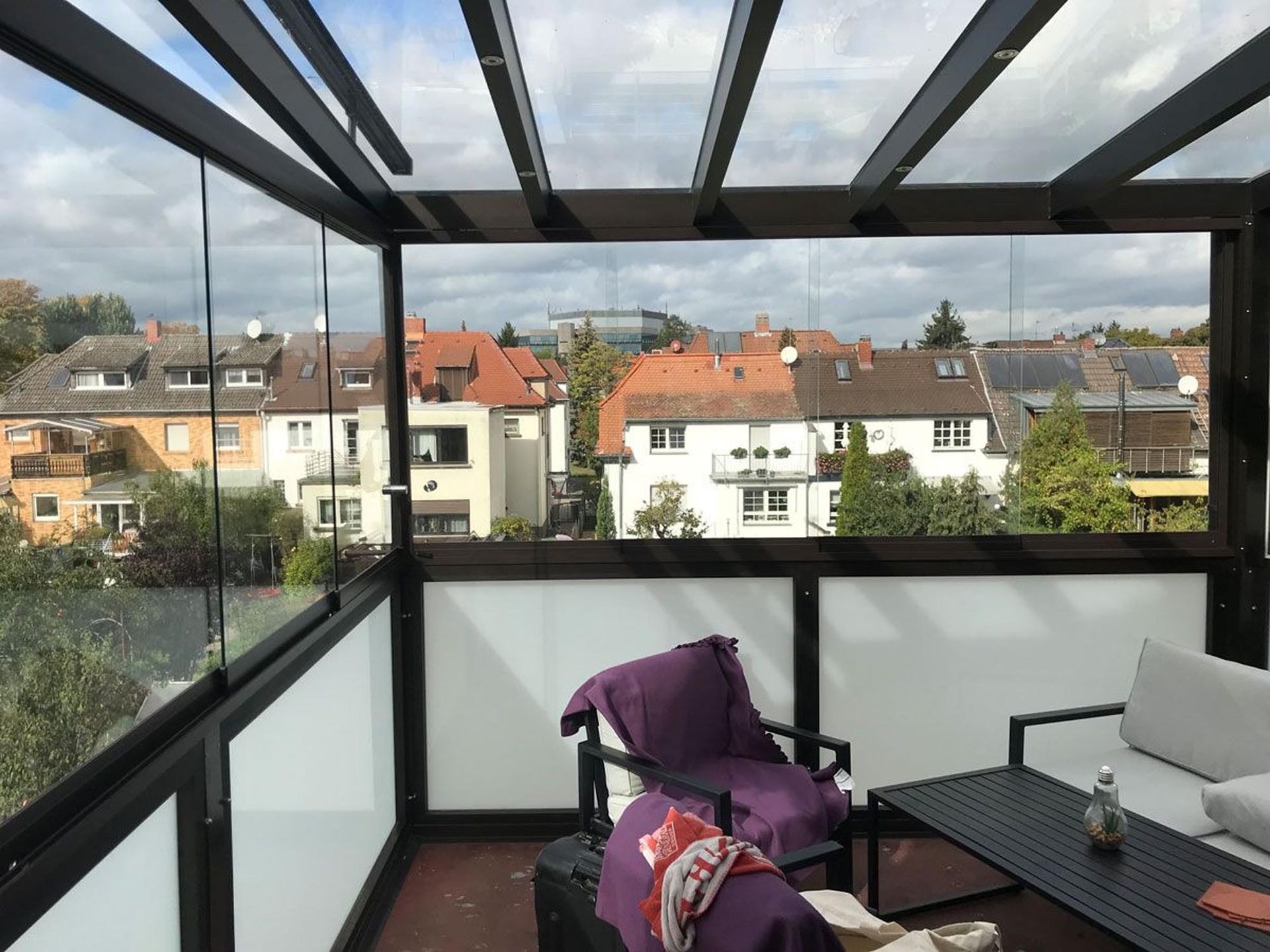Balkon mit Glasgeländer, Terrassenmöbeln und Stadtblick unter einer schwarzen Pergola.